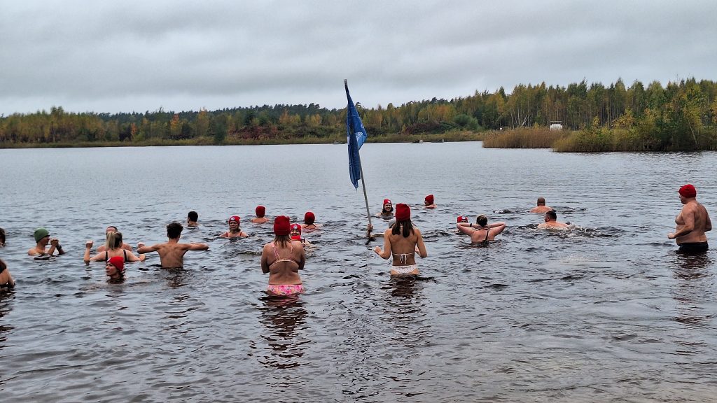 Ziemas peldētāji peldas ūdenskrātuvē, vairākiem galvā sarkanas cepures.