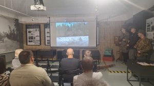 Projekta “Sēlijas Mežabrāļi” prezentācijas dalībnieki skatās video par mežabrāļu vēsturi, bet līdzās ekrānam stāv trīs pasākuma vadītāji.