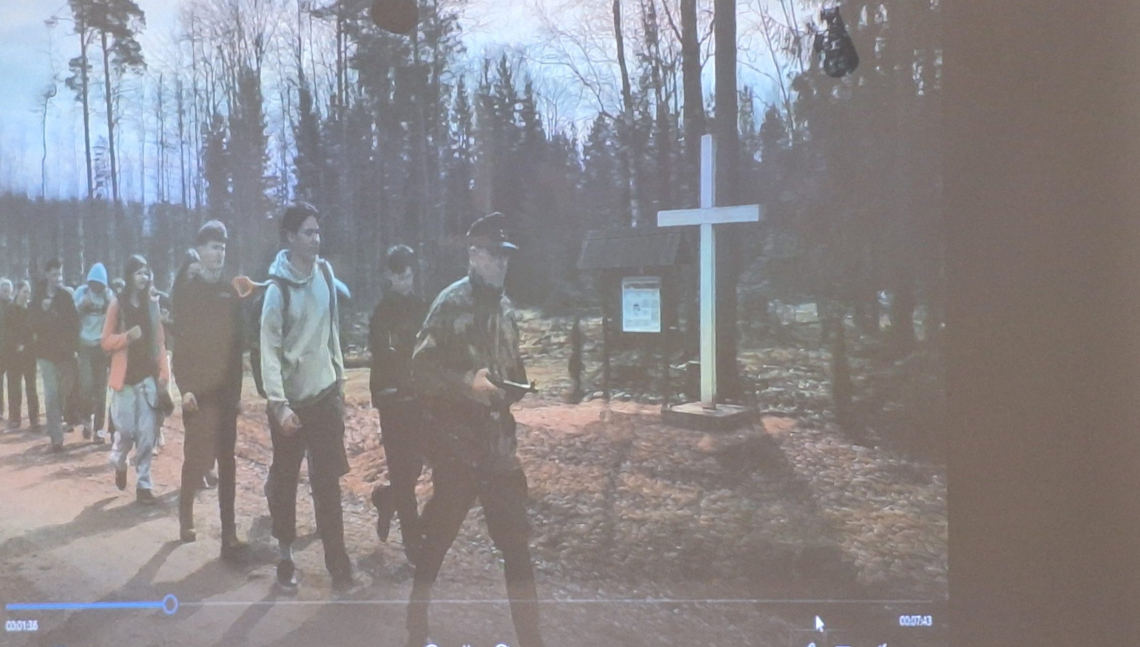 Cilvēku grupa dodas pārgājienā pa meža taku garām baltam krustam, grupas priekšgalā iet cilvēks militārā apģērbā.