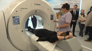 Medicīnas darbiniece demonstrē datortomogrāfu, sievietei guļot uz izmeklējumu galda.