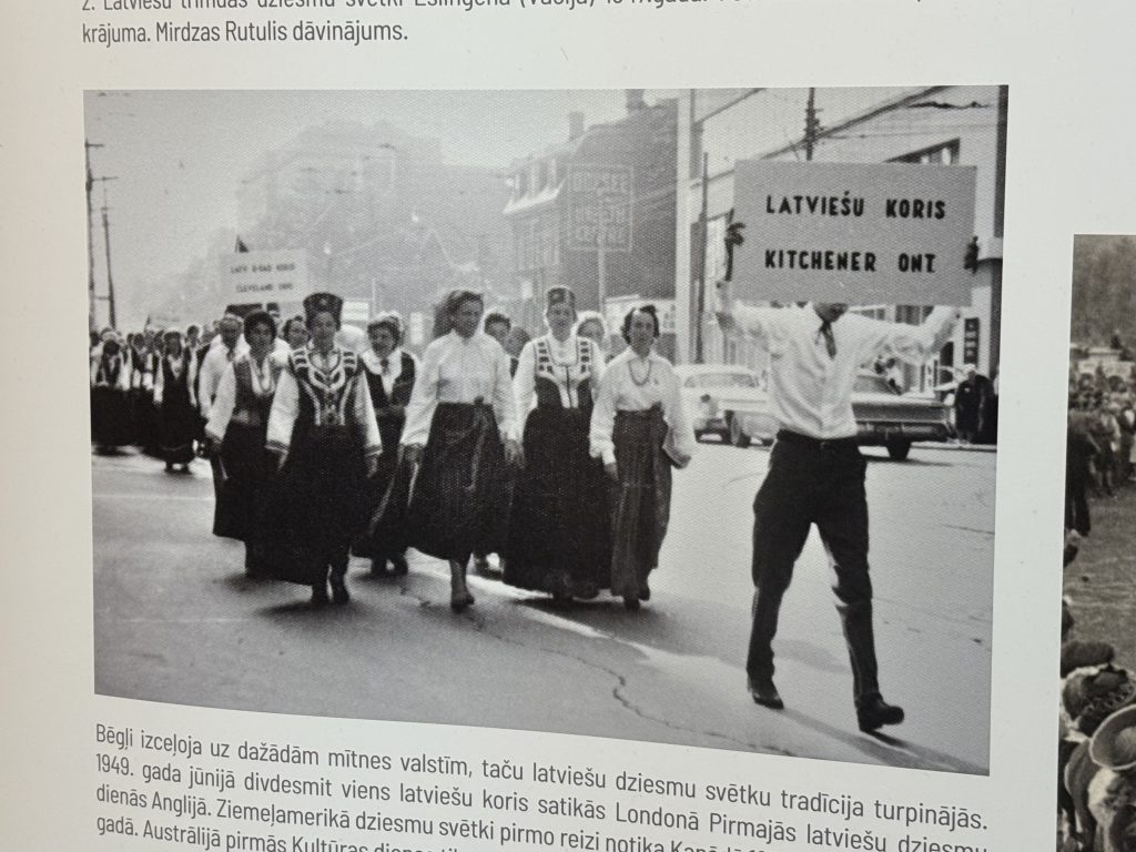 Vēsturiska melnbalta fotogrāfija ar latviešu kori tautastērpos gājienā pa pilsētas ielu.