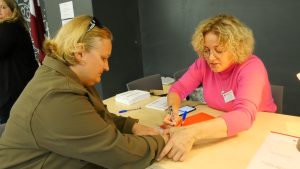 Alt text: Vēlēšanu iecirkņa darbiniece palīdz vēlētājai parakstīties