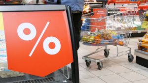 Liela atlaižu zīme ar procentu simbolu pārtikas veikalā, fonā iepirkumu ratiņi ar pārtikas produktiem.