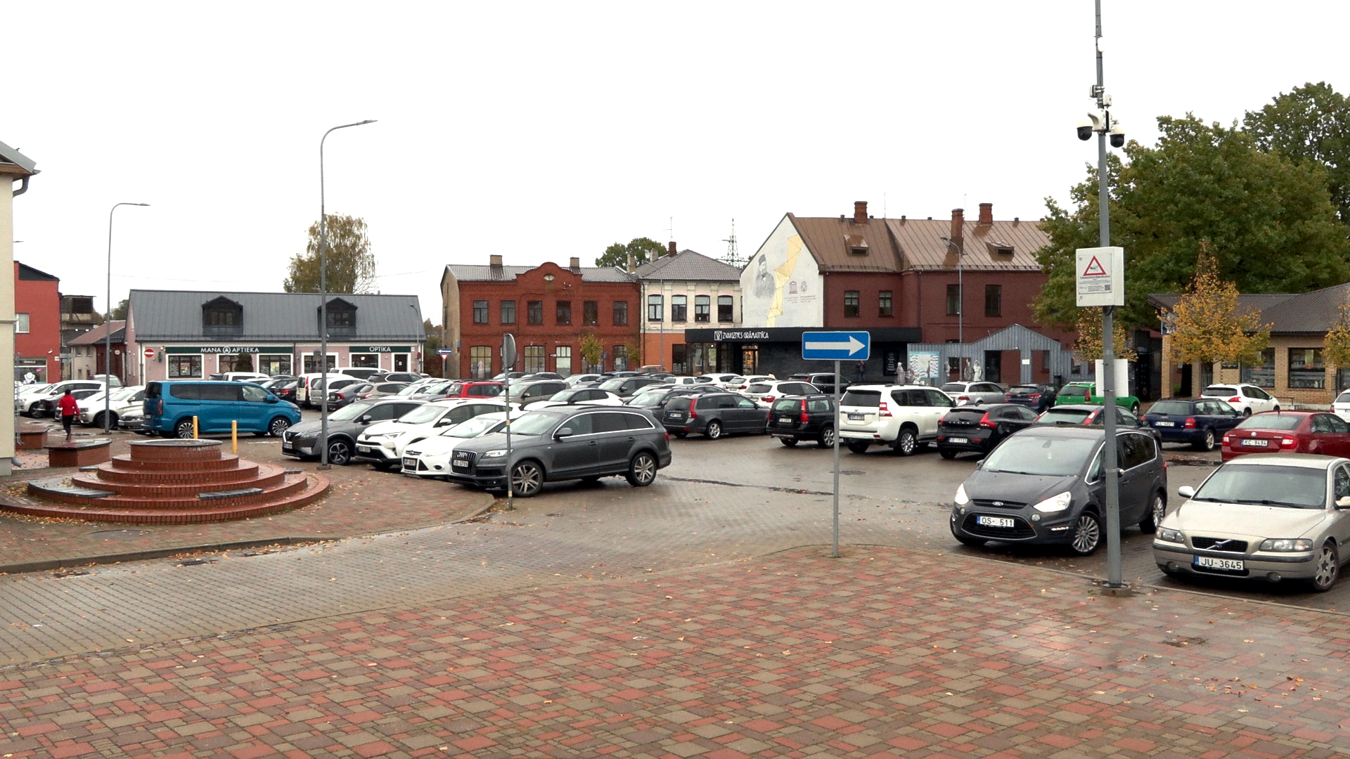 Jēkabpils Vecpilsētas laukums rudenī ar automašīnu stāvlaukumu, apkārtējām ēkām un pilsētas infrastruktūru.
