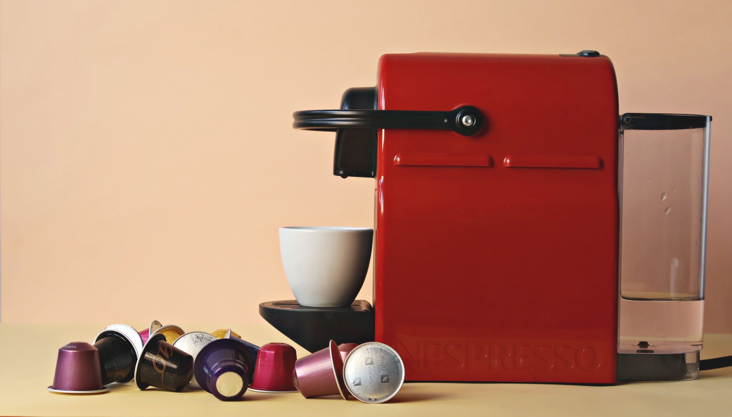 Sarkans Nespresso kapsulu kafijas automāts ar baltu kafijas tasīti un dažādu krāsu kafijas kapsulām uz galda.