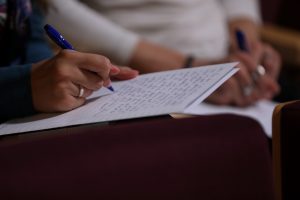 Diktāta dalībnieki ar zilām pildspalvām raksta tekstu uz papīra lapām, pārbaudot savas valodas zināšanas.