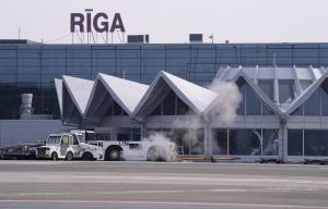 Rīgas lidostas termināļa ēka ar uzrakstu “RĪGA”, priekšplānā lidostas apkalpošanas tehnika un lidlauks.
