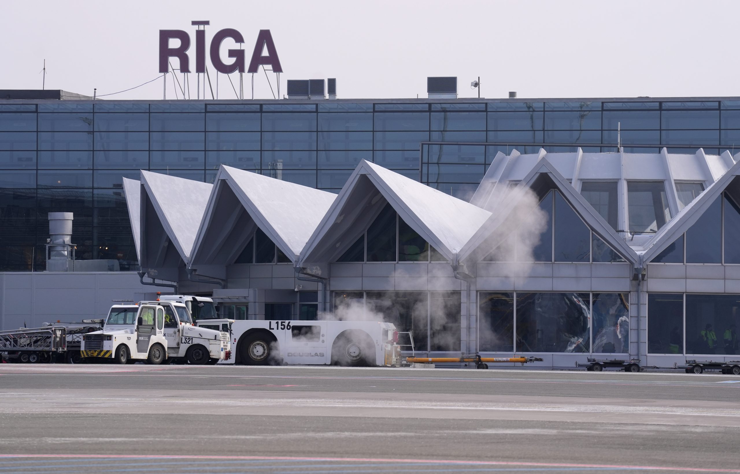 Rīgas lidostas termināļa ēka ar uzrakstu “RĪGA”, priekšplānā lidostas apkalpošanas tehnika un lidlauks.