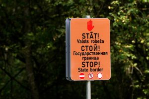 Oranža brīdinājuma zīme pie Latvijas valsts robežas ar uzrakstu “STĀT! Valsts robeža” latviešu, krievu un angļu valodā.