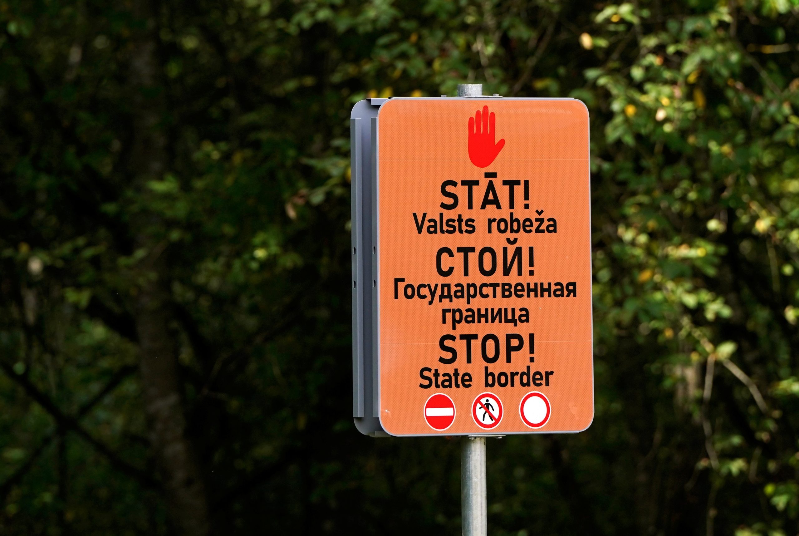 Oranža brīdinājuma zīme pie Latvijas valsts robežas ar uzrakstu “STĀT! Valsts robeža” latviešu, krievu un angļu valodā.