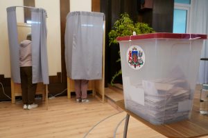 Vēlēšanu urna ar balsu aploksnēm un vēlētāji balsošanas kabīnēs
