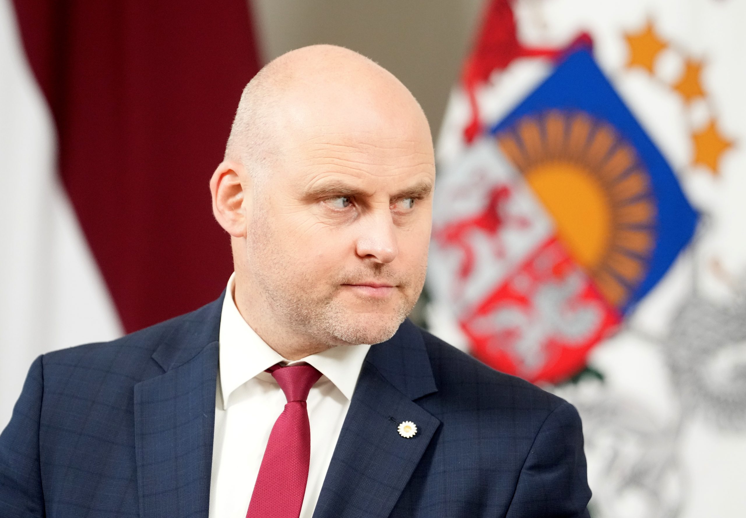 Ministru prezidents Andris Kulbergs tumši zilā uzvalkā un sarkanā kaklasaitē, fonā Latvijas karogs un valsts ģerbonis.