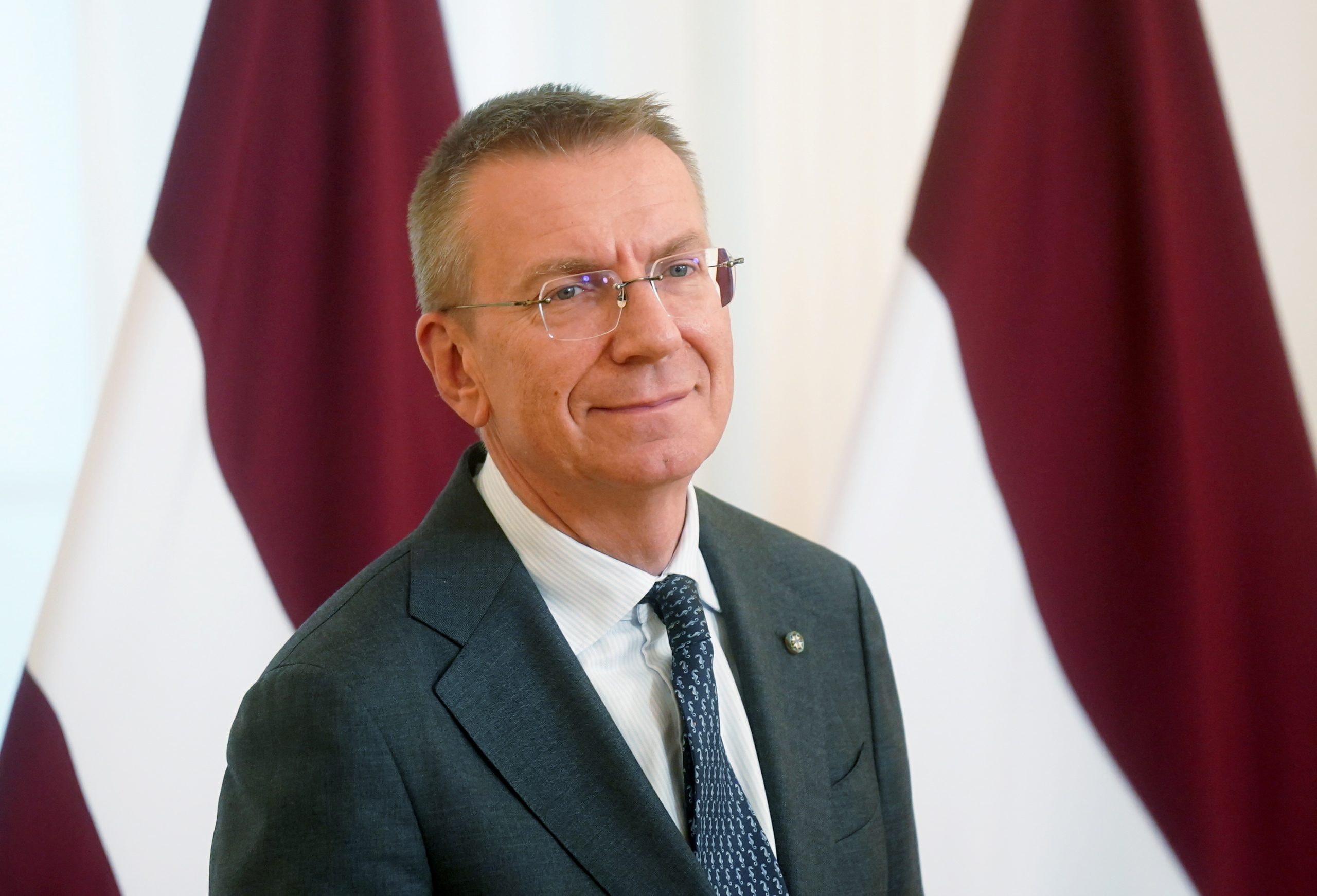 Valsts prezidents Edgars Rinkēvičs tumšā uzvalkā un brillēs, fonā Latvijas valsts karogi.