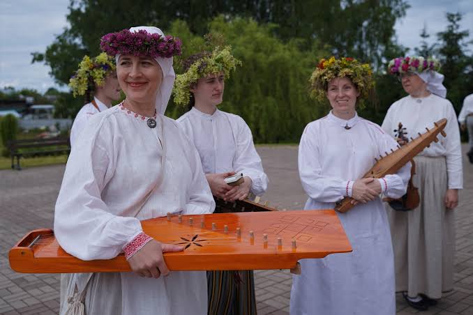 Sievietes tautastērpos un ziedu vainagos ar tradicionālajiem mūzikas instrumentiem.