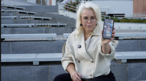 Sieviete ar brillēm rāda viedtālruņa ekrānu ar ziņu saturu