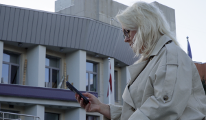 Sieviete ar viedtālruni pie ēkas, kur redzams Latvijas karogs