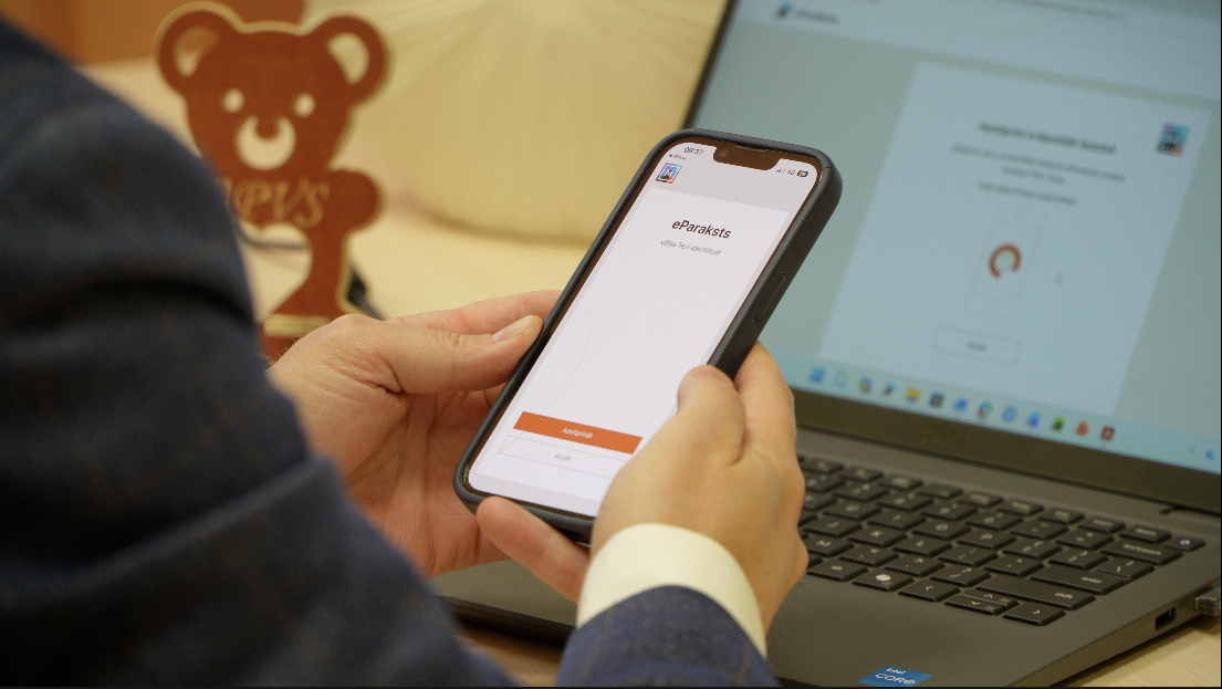 Persona izmanto eParaksts mobile lietotni viedtālrunī, lai elektroniski parakstītu dokumentu datorā.