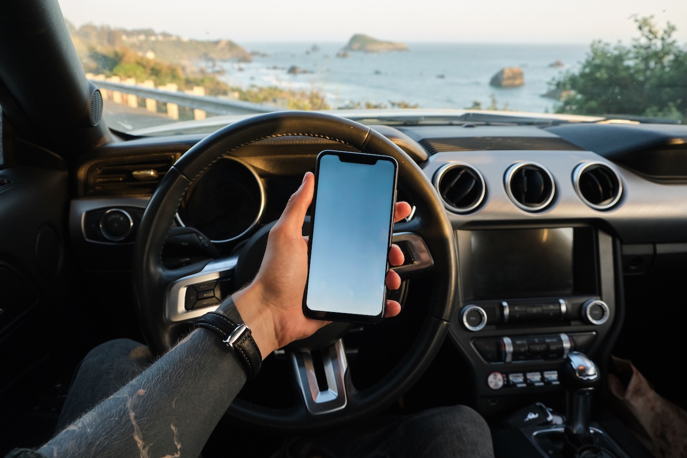 Autovadītājs tur rokā mobilo telefonu pie automašīnas stūres