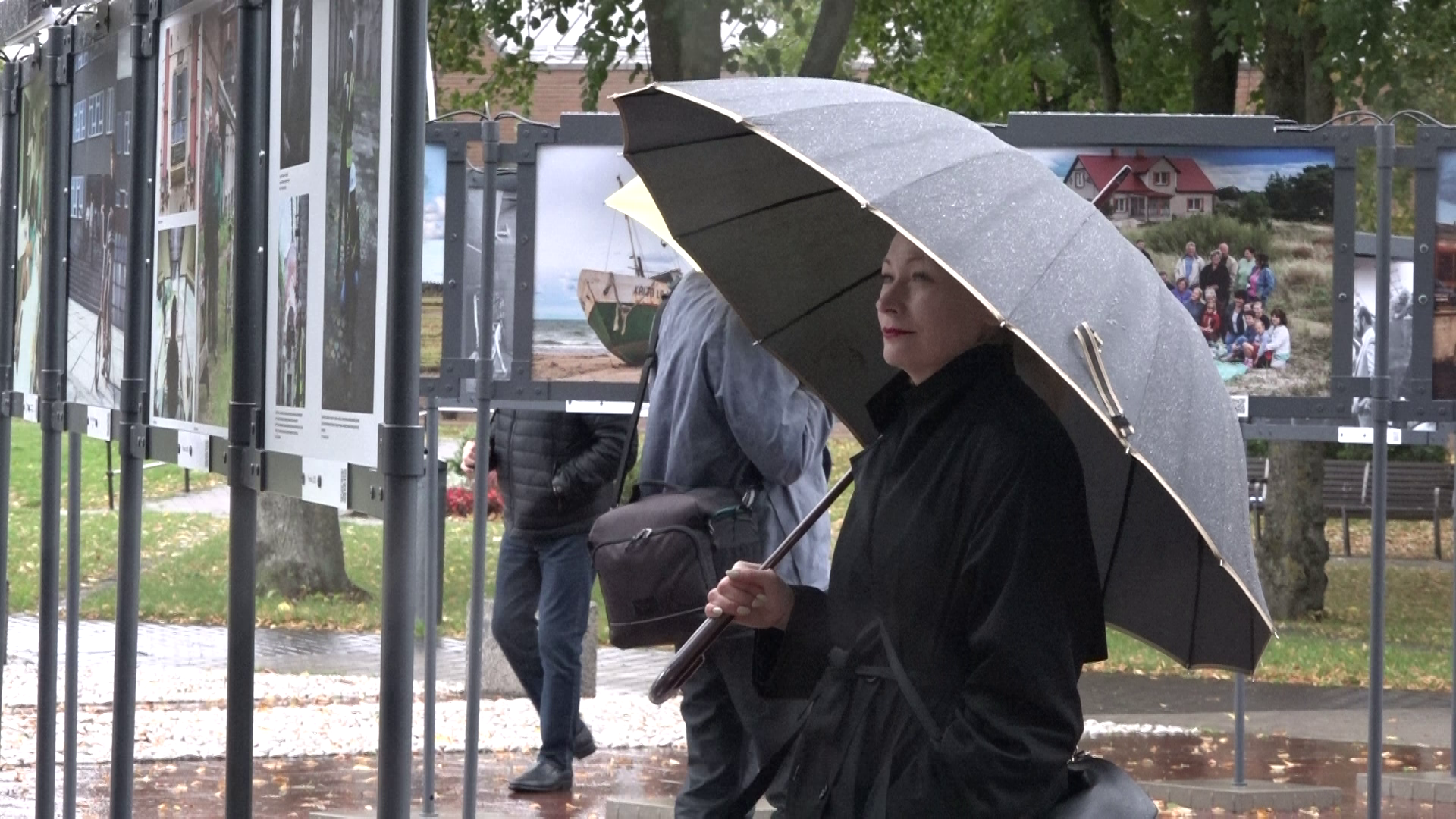 Sieviete ar lietussargu aplūko brīvdabas fotogrāfiju izstādi Liepājā.