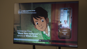 Liepājas okupācijas muzeja ekrānā redzams animācijas filmas “Mana tēva noslēpumi” plakāts un paziņojums par tikšanos ar autoru Mišelu Kišku.